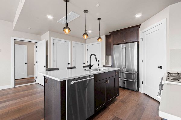 A modern kitchen features a central island with a sink, pendant lighting, stainless steel appliances, and wooden floors, creating a sleek design.