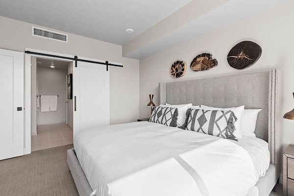 A modern bedroom with a large bed, decorative pillows, wall art, and a sliding door leading to a bathroom with towels hanging.