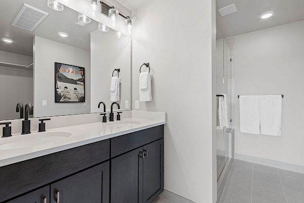 A modern bathroom has a double sink vanity with black fixtures, a large mirror, towels, a framed picture, and a glass-enclosed shower.
