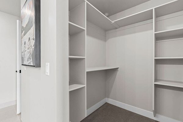 Image of an empty walk-in closet featuring built-in shelving, hanging rods, and a light grey color scheme. A section of the adjacent room is visible.