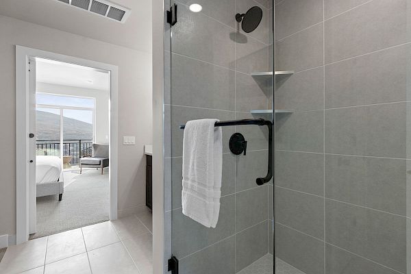 The image shows a modern bathroom with a glass-enclosed shower, a white towel on a black towel bar, and an adjacent room with a bed in the background.