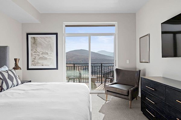 A modern bedroom with a bed, an armchair, dresser, wall art, and a TV. A large window and balcony offer a scenic mountain view in the background.