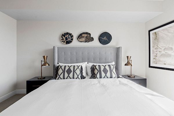 A modern bedroom with a large bed, gray headboard, geometric pillows, and wall art above the bed. There are two bedside tables with gold lamps.