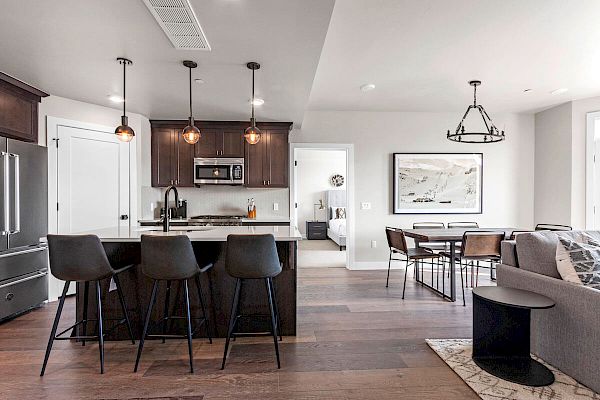 A modern open-concept kitchen and living area with dark cabinetry, an island with barstools, a dining table, and a sofa facing a framed artwork.