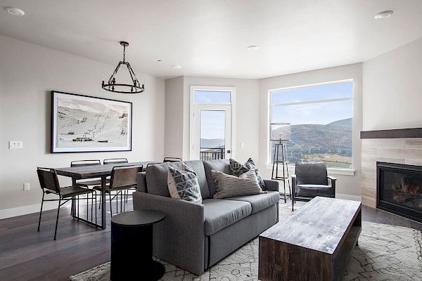 A modern living room with a gray sofa, dining table, fireplace, and large windows offering a scenic view of mountains.