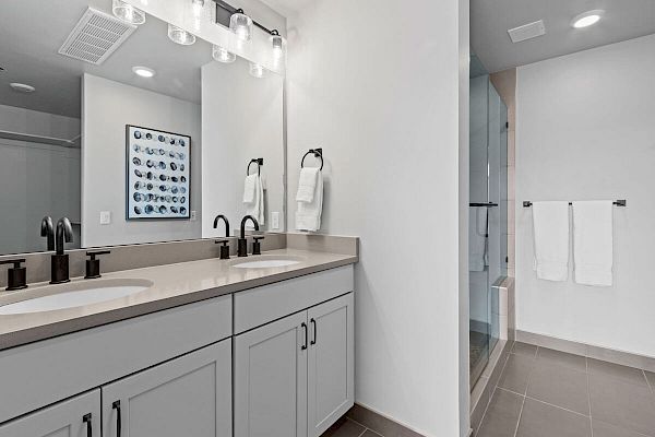 A modern bathroom with double sinks, a large mirror, two black faucets, a glass shower, and white cabinetry. Towel racks hold white towels.