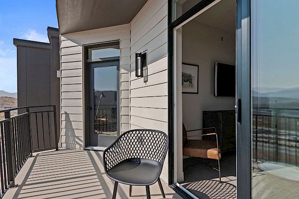 A modern balcony with a black mesh chair overlooks a scenic view. Inside, there's a sliding glass door leading to a room with a chair and a TV.