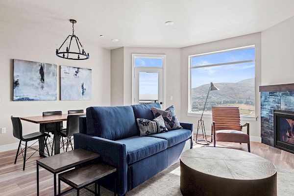 A modern living room with a blue sofa, large window, fireplace, wall art, dining table, and chairs, overlooking scenic hills, ending the sentence.