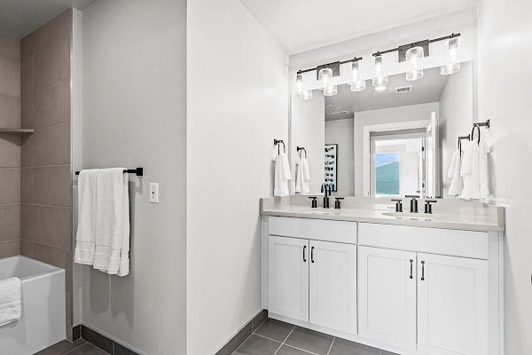 A modern bathroom features a double sink with a large mirror, light fixtures, and white cabinets. A bathtub with towels is visible to the left.