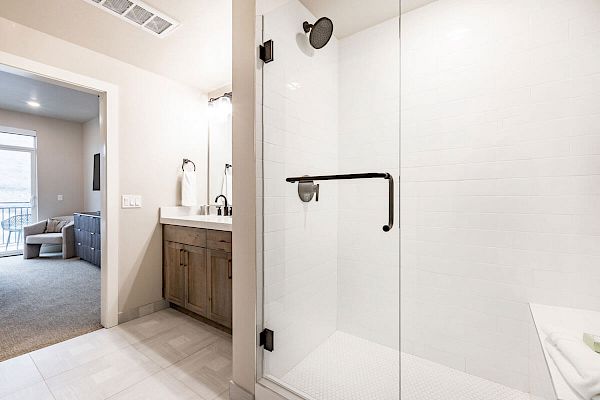 The image shows a modern bathroom with a glass shower enclosure, a wooden vanity with a sink, and a view into an adjacent carpeted room.