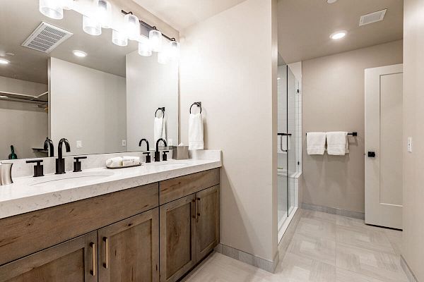 This image shows a modern bathroom with double sinks, a mirror with lights, wooden cabinets, towels, and a shower in the background.