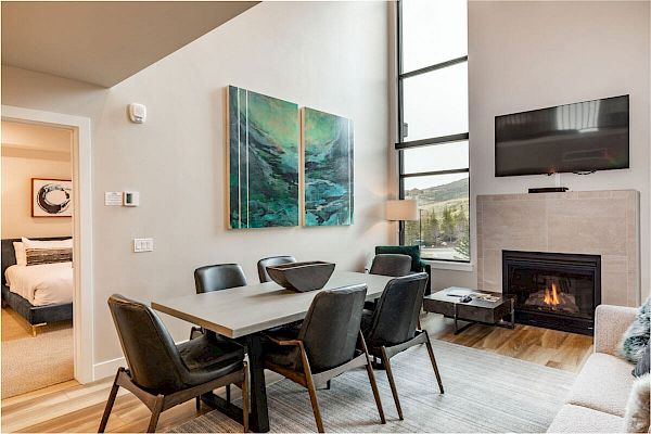A cozy, modern living-dining room with a wall-mounted TV, fireplace, dining table, abstract art, and large windows. Adjacent bedroom visible.