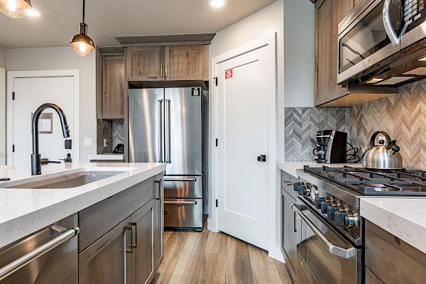 A modern kitchen with stainless steel appliances, wooden cabinets, white countertops, and a herringbone backsplash.