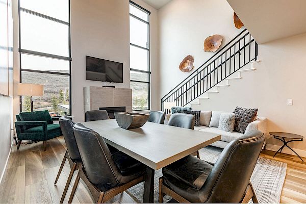 A modern living and dining area with a wooden table, black chairs, a sectional sofa, large windows, wall decor, and a staircase.
