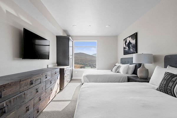 A modern bedroom with two beds, a wooden dresser, a wall-mounted TV, a nightstand with a lamp, and a window with a mountain view ends the sentence.
