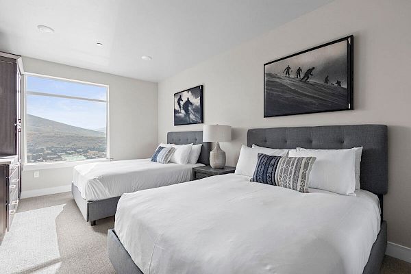 A modern bedroom with two beds, grey headboards, white bedding, a large window, and ski-themed artwork above the beds, creating a cozy vibe.