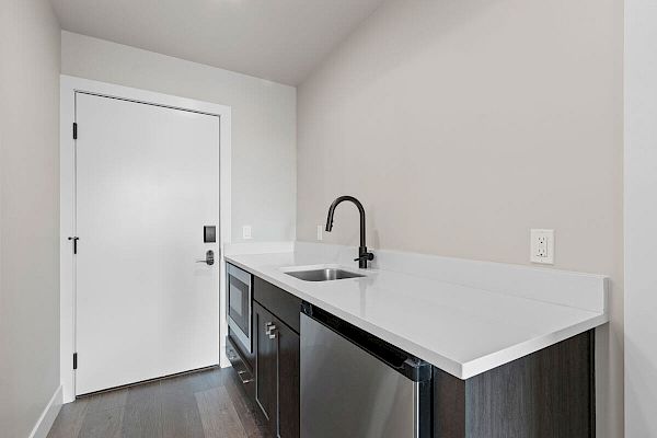 A small, modern kitchenette with a sink, dark cabinets, a white countertop, and a door in the background.