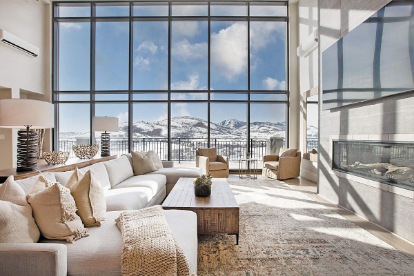 A modern living room with large windows showcasing a snowy mountain view, furnished with a sectional sofa, chairs, and a cozy fireplace.