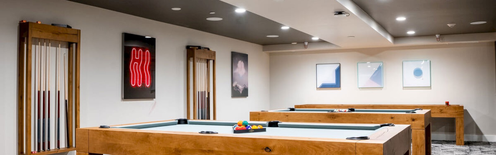The image shows a game room with pool tables, wall art, cue racks, and modern lighting, featuring a patterned carpet and neatly arranged decor.