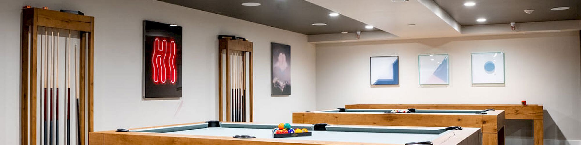 The image shows a game room with pool tables, wall art, cue racks, and modern lighting, featuring a patterned carpet and neatly arranged decor.