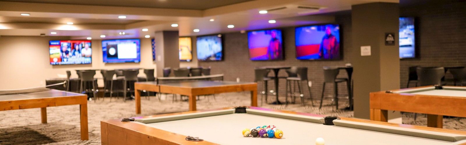 The image shows a modern game room with pool tables, chairs, tables, and multiple TV screens on the walls.