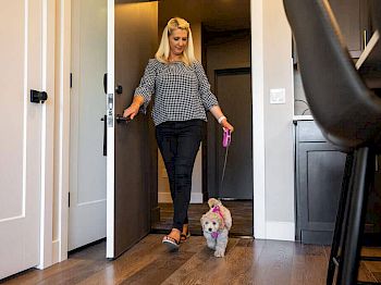 A woman is walking out of a room with a small dog on a leash, inside a house with wooden flooring and modern decor, ending the sentence.