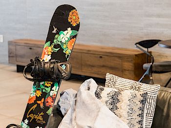 A snowboard with floral designs is propped up indoors, resting against a couch with pillows and a blanket.