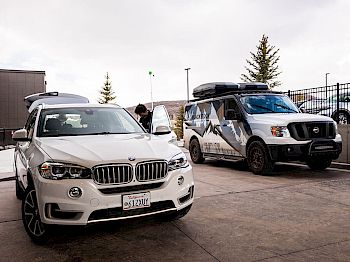 Image features a white BMW SUV and a white van with a mountain graphic in an outdoor setting; two people nearby.
