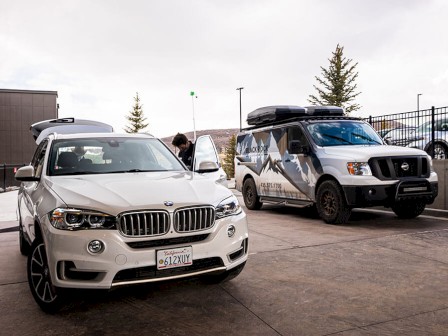 Image features a white BMW SUV and a white van with a mountain graphic in an outdoor setting; two people nearby.