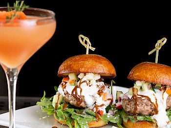 The image shows two sliders with garnishes on a plate and a cocktail next to them, all set against a dark background.