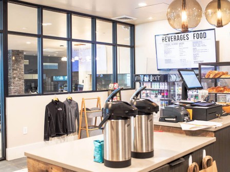 The image shows a modern café interior with beverage dispensers, a menu board, pastries, shelves with items, and a seating area with glass walls.