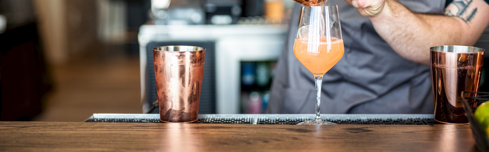 A bartender pours a pink cocktail from a shaker into a glass. Copper shakers and a cocktail setup are on the wooden bar counter.