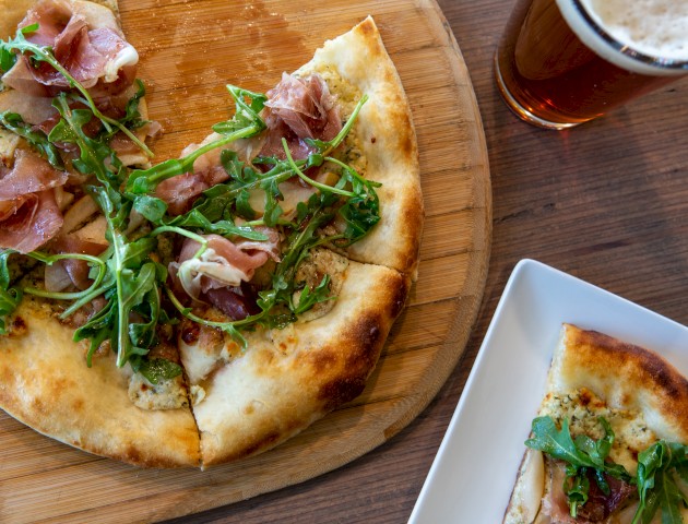The image shows a pizza topped with prosciutto, arugula, and cheese on a wooden board, alongside a glass of beer and a small pizza slice.