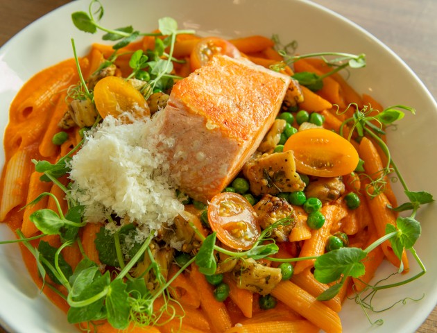 A dish with pasta, topped with grilled salmon, grated cheese, peas, cherry tomatoes, and herbs, all garnished with sprouts.