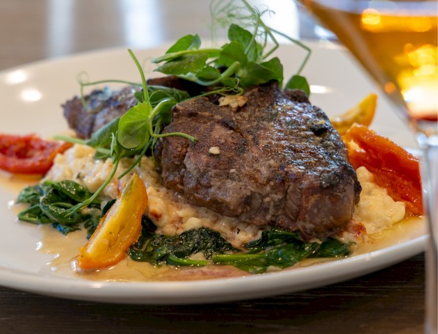 The image shows a plated dish with a grilled steak, colorful vegetables, greens, and a glass of wine on a wooden table.