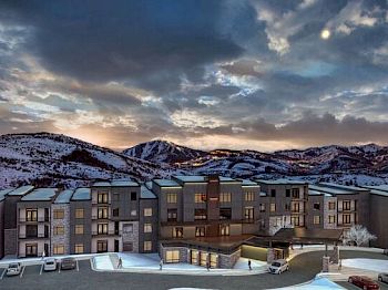 The image shows a modern apartment complex set against a backdrop of snow-covered mountains under a cloudy evening sky, with parked cars in front.