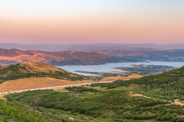 A scenic landscape with rolling green hills, a winding road, and a calm lake under a clear, pink-hued sky at sunset.