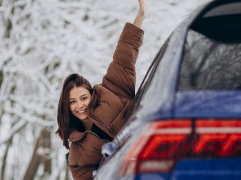 A person in a brown jacket leans out of a blue car's window, smiling and waving their hand, against a snowy forest background.