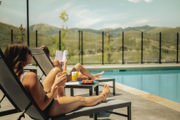 People relax by a pool on lounge chairs, with one reading a book, drinking, and a plate of snacks nearby on a sunny day.