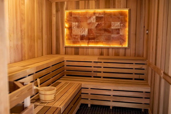 The image shows a wooden sauna with bench seating, a bucket and ladle, and a backlit salt wall panel, creating a warm ambiance.