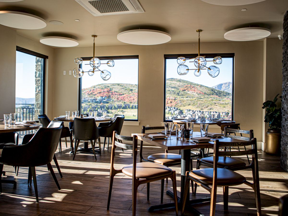 A modern restaurant with large windows showing a scenic mountain view, elegant seating, wooden floors, and stylish light fixtures.