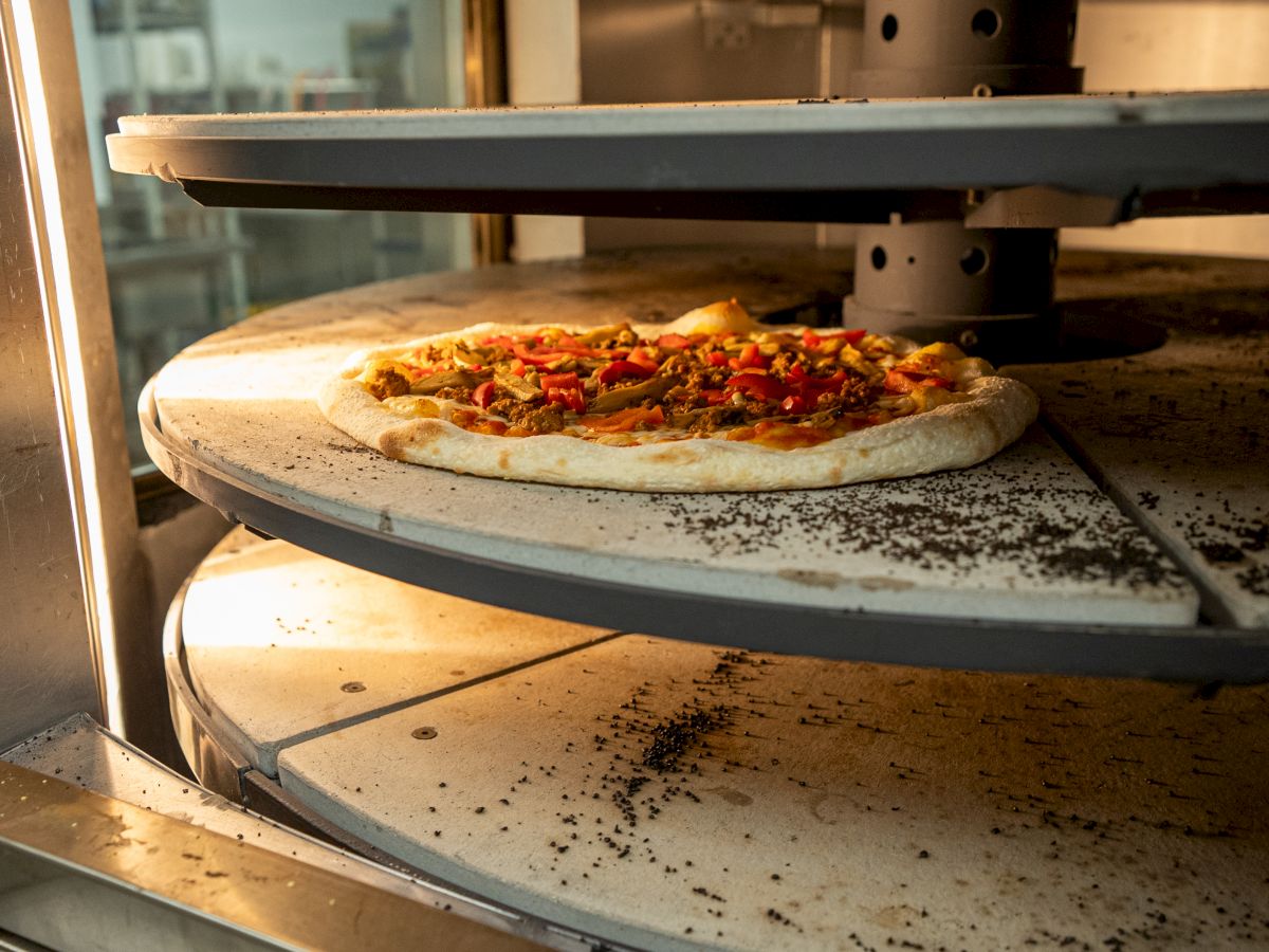 A pizza is baking on a rotating stone deck inside an oven. The stone surface shows signs of previous use, with some residue visible.
