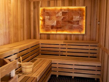 The image shows the interior of a wooden sauna with benches, a bucket, and a ladle, featuring a glowing panel on the wall.