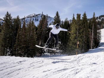 A person in a white jacket is performing a jump on a snowboard in a snowy, forested mountainous area on a clear day.