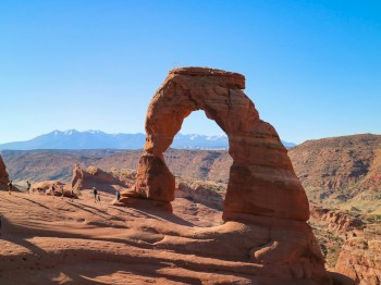 The image shows Delicate Arch in Arches National Park, Utah, with a clear blue sky and distant mountains. There are some people around the arch.