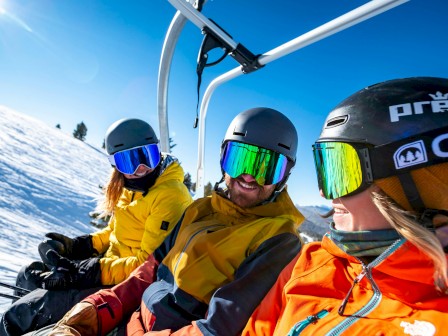 Three people in ski gear, including helmets and goggles, sitting on a ski lift with snow-covered mountains in the background and a bright blue sky.