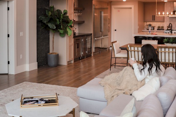 A bright, modern living area with a white sectional sofa, glass railing balcony, wooden floors, a round coffee table, and a kitchen bar in the background. End.