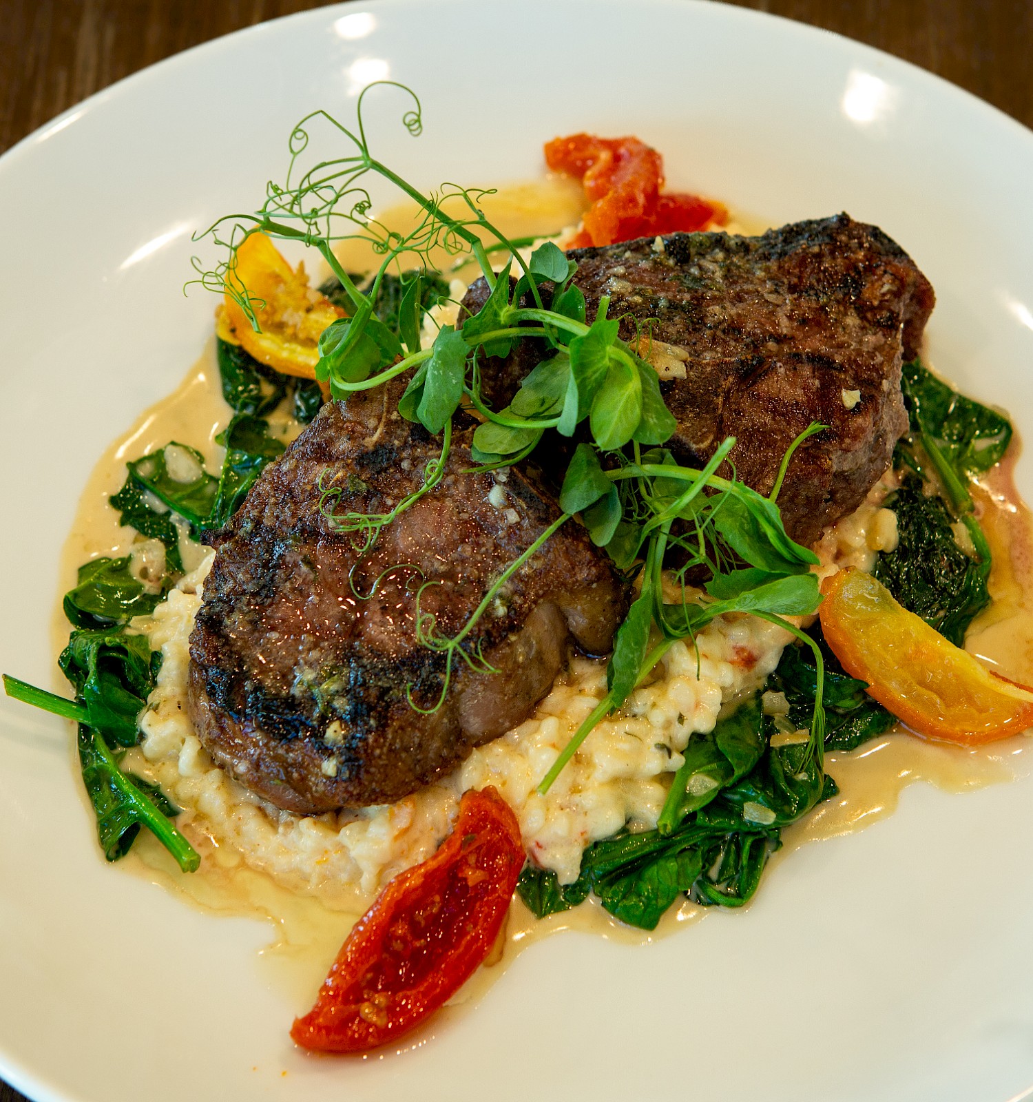 The image shows a plated dish with grilled steak on a bed of risotto, garnished with greens, baby tomatoes, and herbs on a white plate.