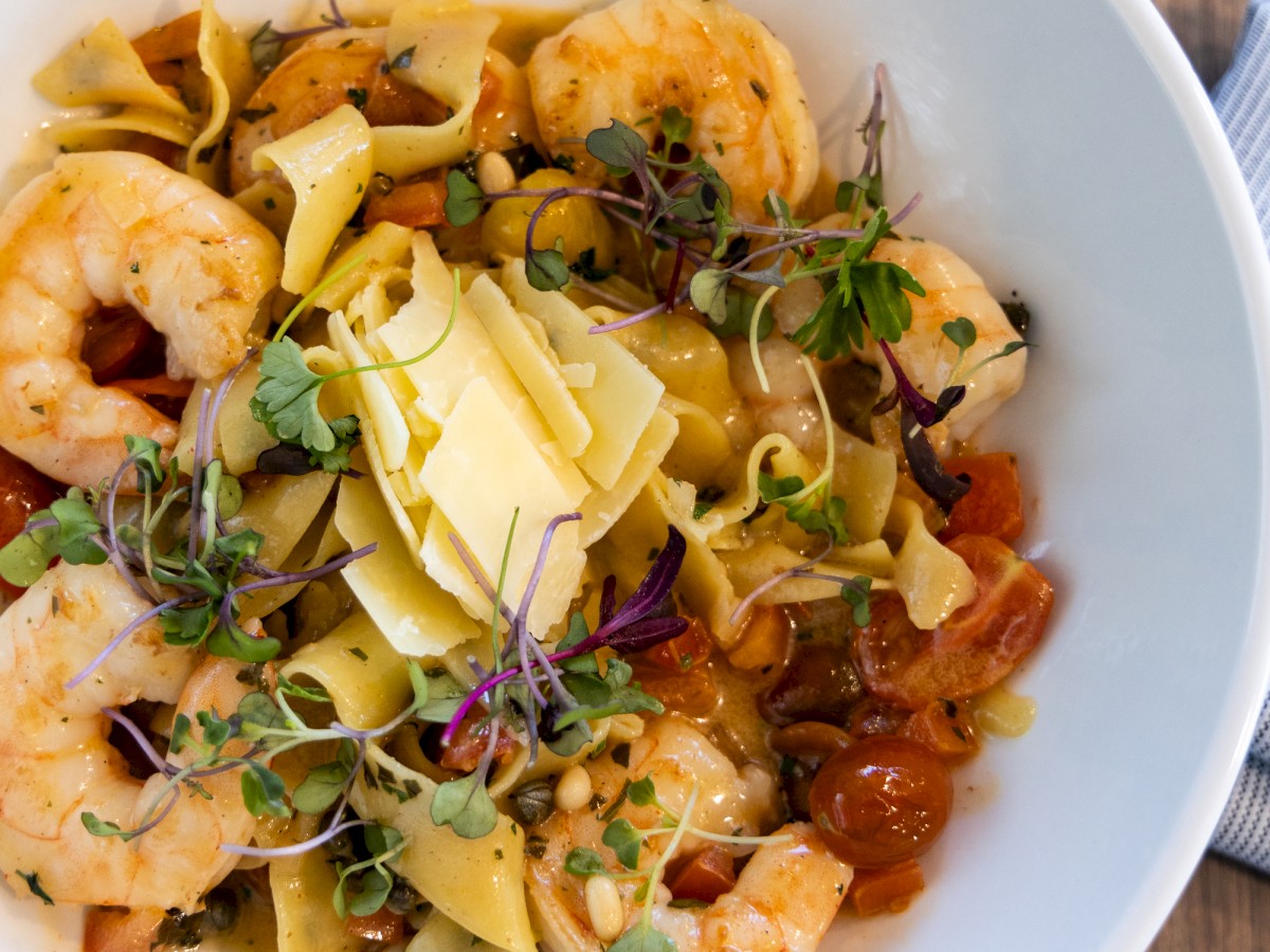 A bowl of pasta topped with shrimp, microgreens, cherry tomatoes, and cheese slices on a wooden surface with a napkin beside it.