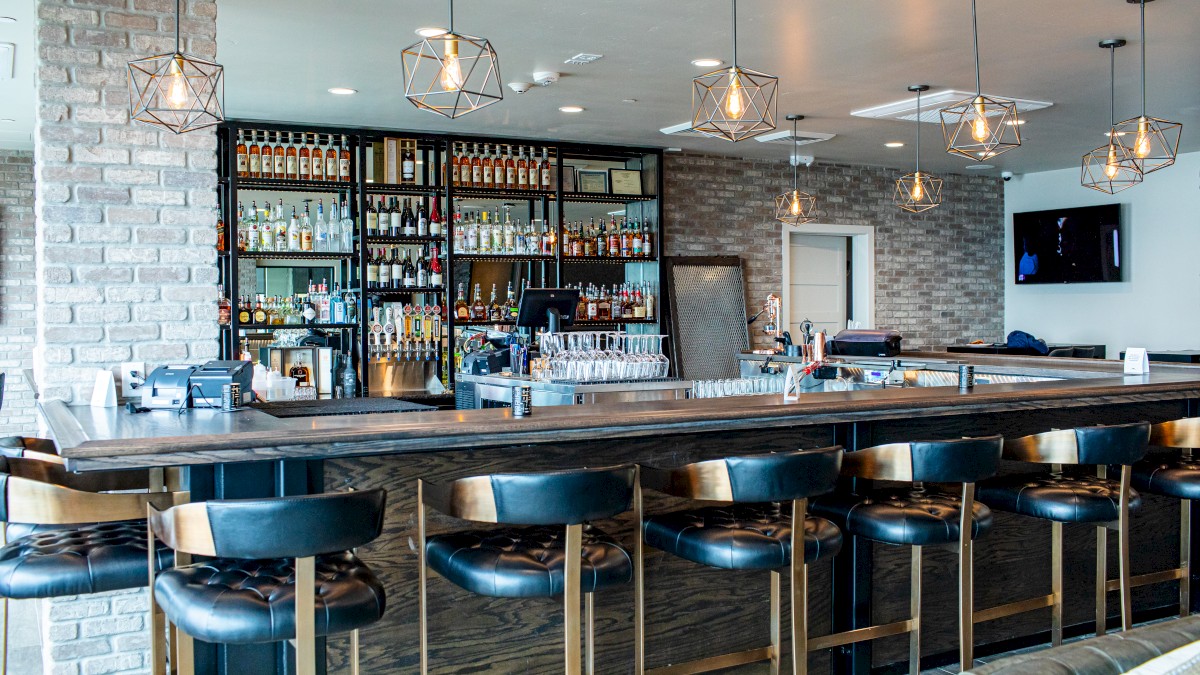 A modern bar with a wooden counter, black leather stools, a brick wall, and shelves stocked with various bottles of alcohol.
